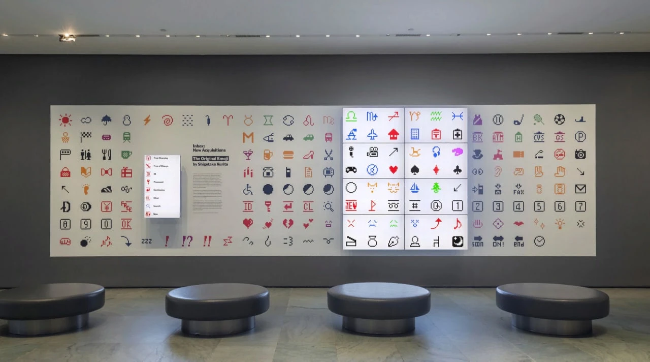 Збільшені емодзі на світлому панно виставки MoMA; перед стіною стоять чотири темні круглі сидіння.