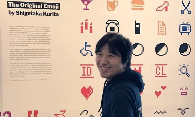 Сіґетака Куріта усміхається перед виставковим панно з написом The Original Emoji та збільшеними піктограмами.