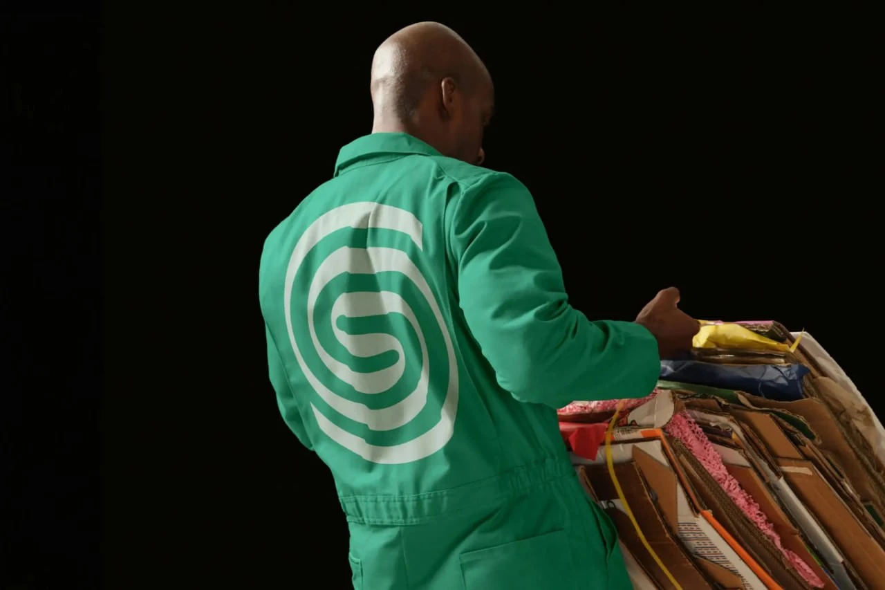 A person wearing a green work jacket with a large white Sustana symbol on the back, beside stacks of paper.